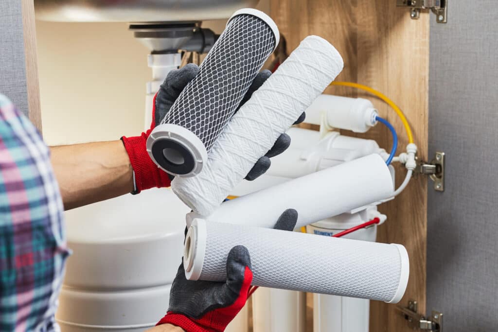 A plumber's hands holding a cartridge to replace a water filter in the kitchen. Fixing the water filtration purification osmosis system.